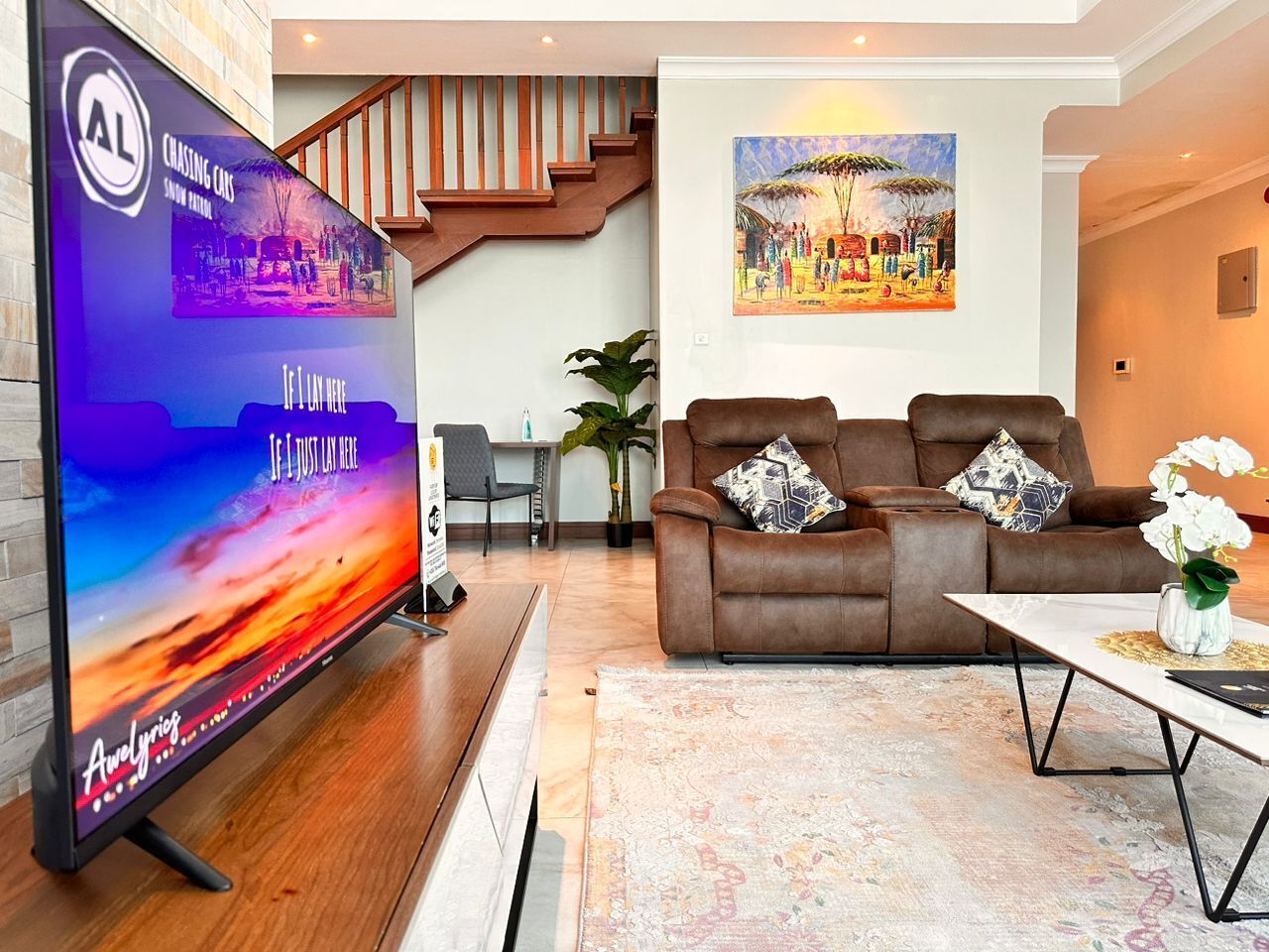 Ein modernes Wohnzimmer mit einem großen Flachbildfernseher, einem braunen Ledersofa mit gemusterten Kissen, einem Couchtisch mit einer Vase weißer Blumen und einem Bild an der Wand.