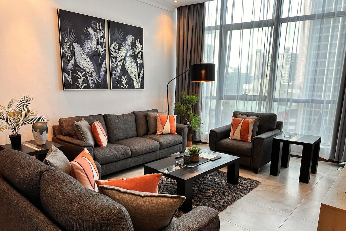 Een moderne woonkamer met een grijze bank, een kleinere fauteuil, een salontafel en een vloerlamp. Er zijn twee grote ramen met dunne gordijnen, en een schilderij van papegaaien aan de muur.