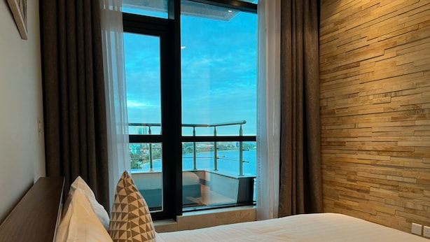 A hotel room with a bed, a view of the sea through a balcony, and a wooden wall. - Fairview Luxury Apartments (Dar es Salaam)