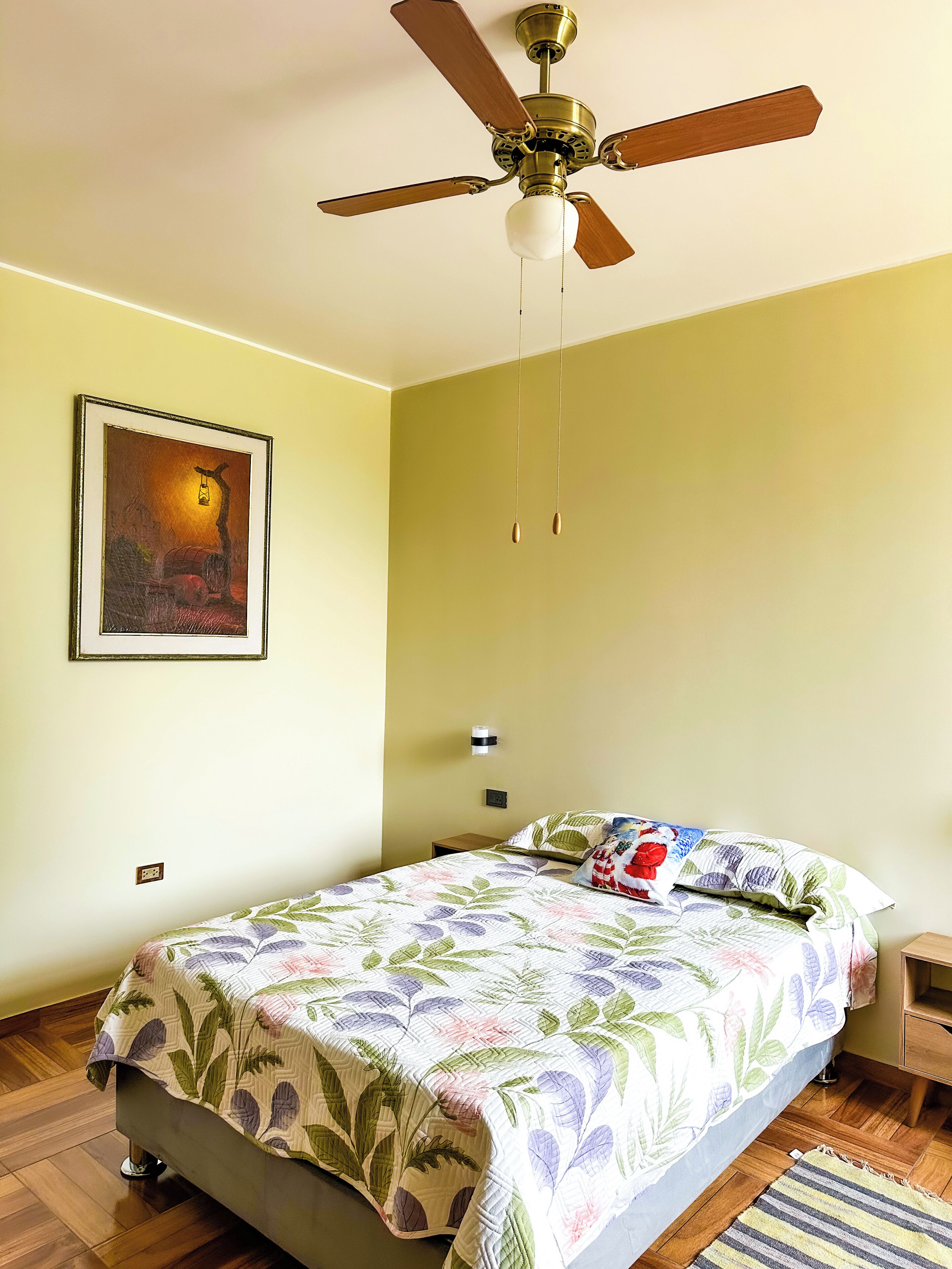 Un dormitorio con cama, ventilador de techo, lámpara de pared, un cuadro enmarcado y una mesita de noche.