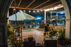 Un patio al aire libre con mesas y sillas, rodeado de plantas en macetas y un gran paraguas.