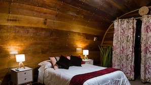 A wooden cabin bedroom with a bed, two bedside tables with lamps, a clock, and a floral curtain. - Glamping Toneles y Viñedos (San Juan Bautista)
