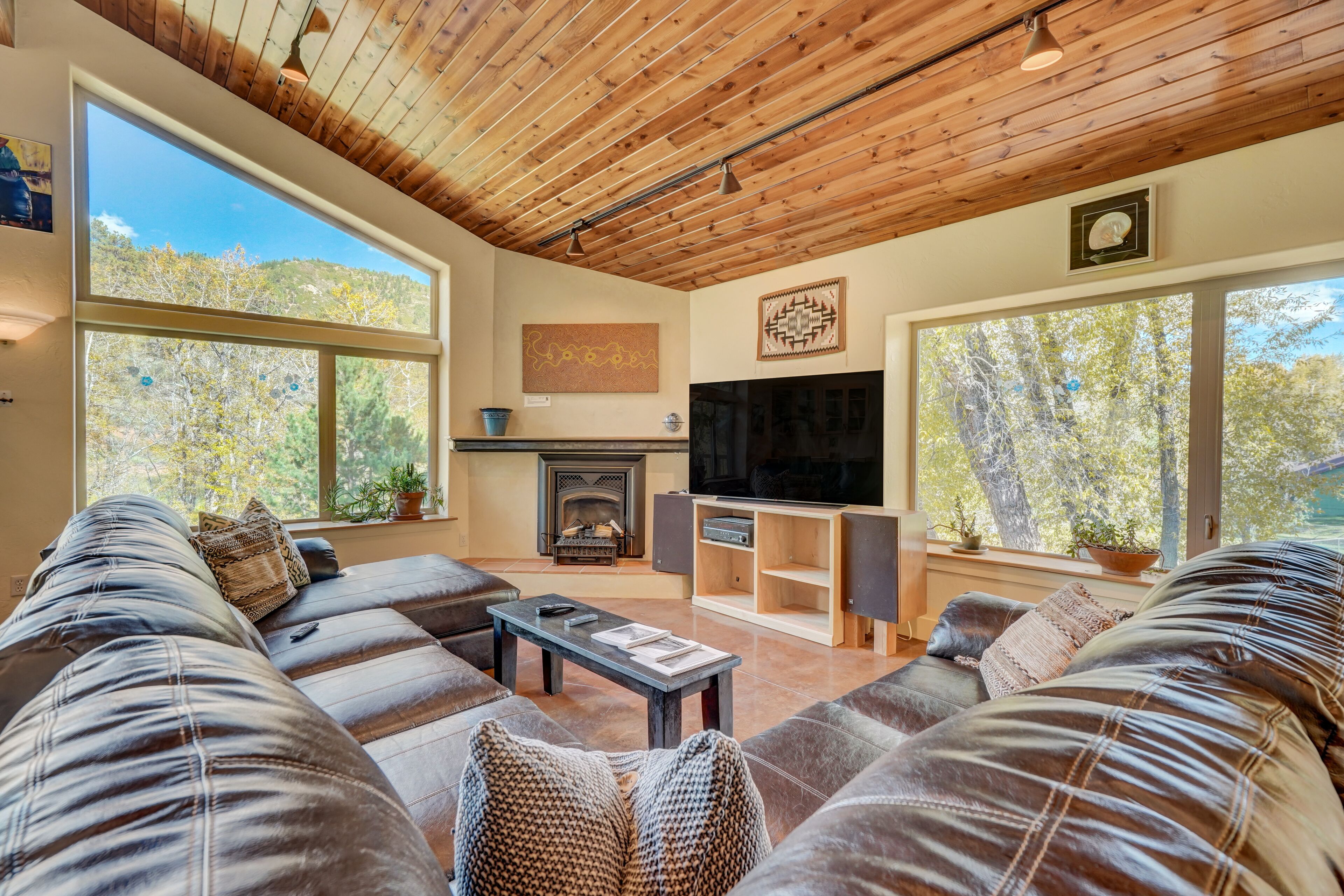 A modern living room with a large sectional sofa, a fireplace, a TV, and large windows offering a view of trees.