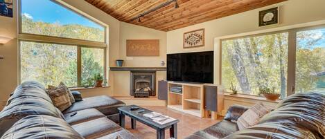 A modern living room with a large sectional sofa, a fireplace, a TV, and large windows offering a view of trees.