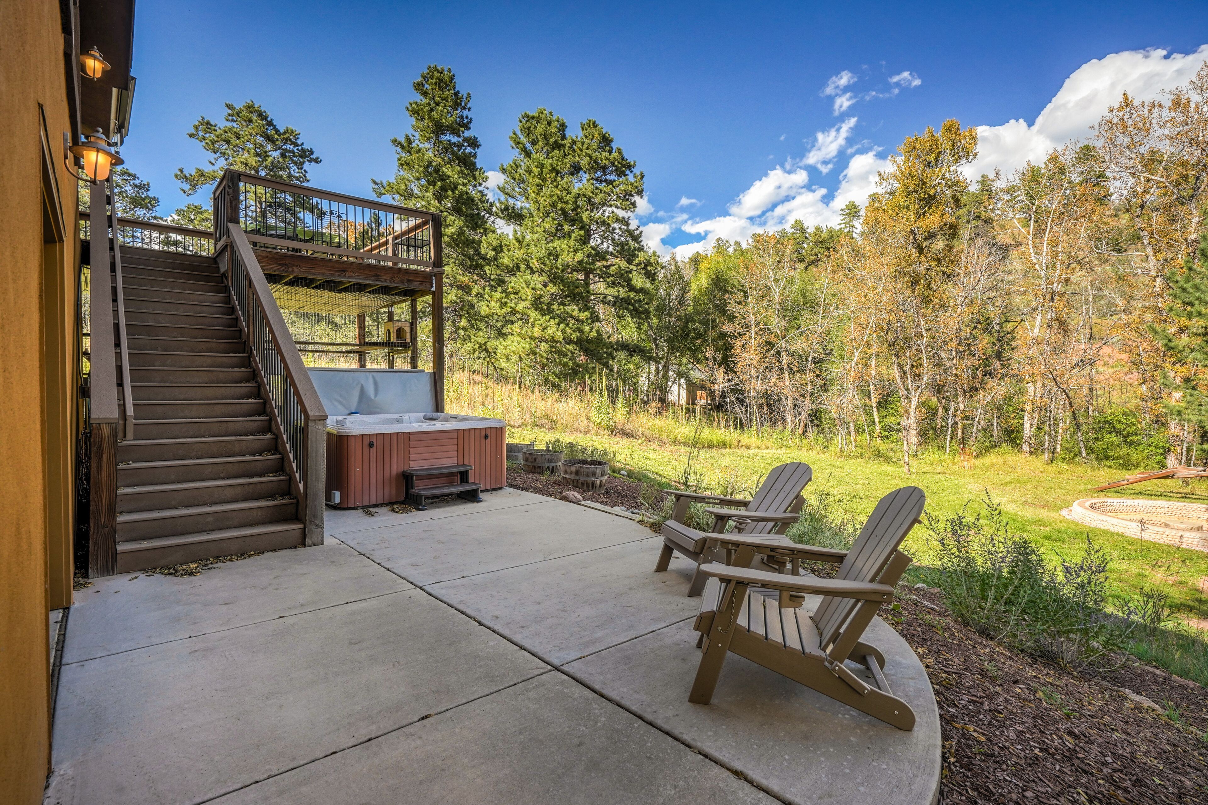A wooden deck with a hot tub, Adirondack chairs, and a staircase leading to a balcony.