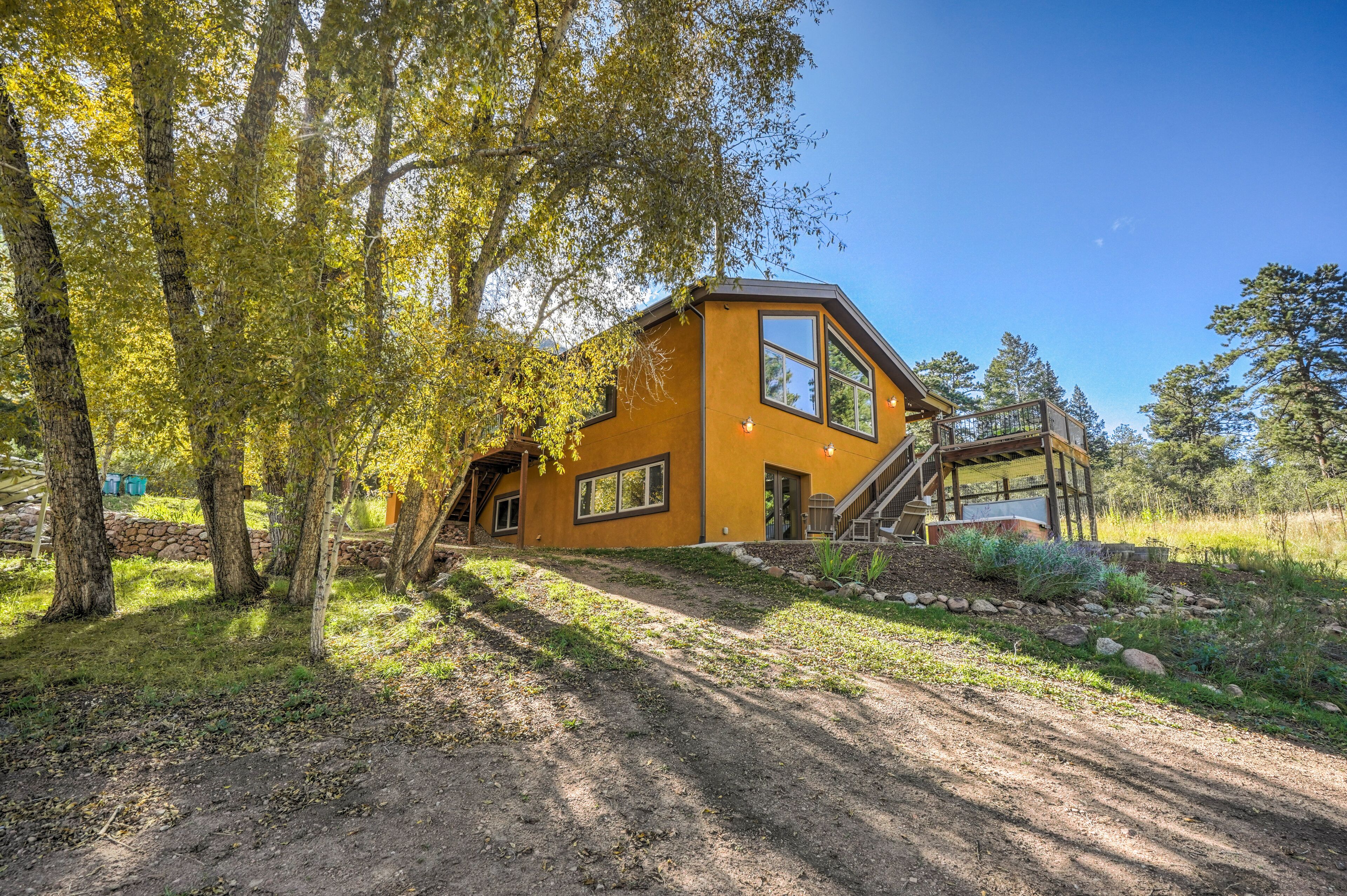 A modern house with a large deck, surrounded by trees and a dirt path.
