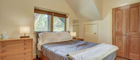A bedroom with a bed, a wooden dresser, a nightstand, a window with blinds, and a wooden door.