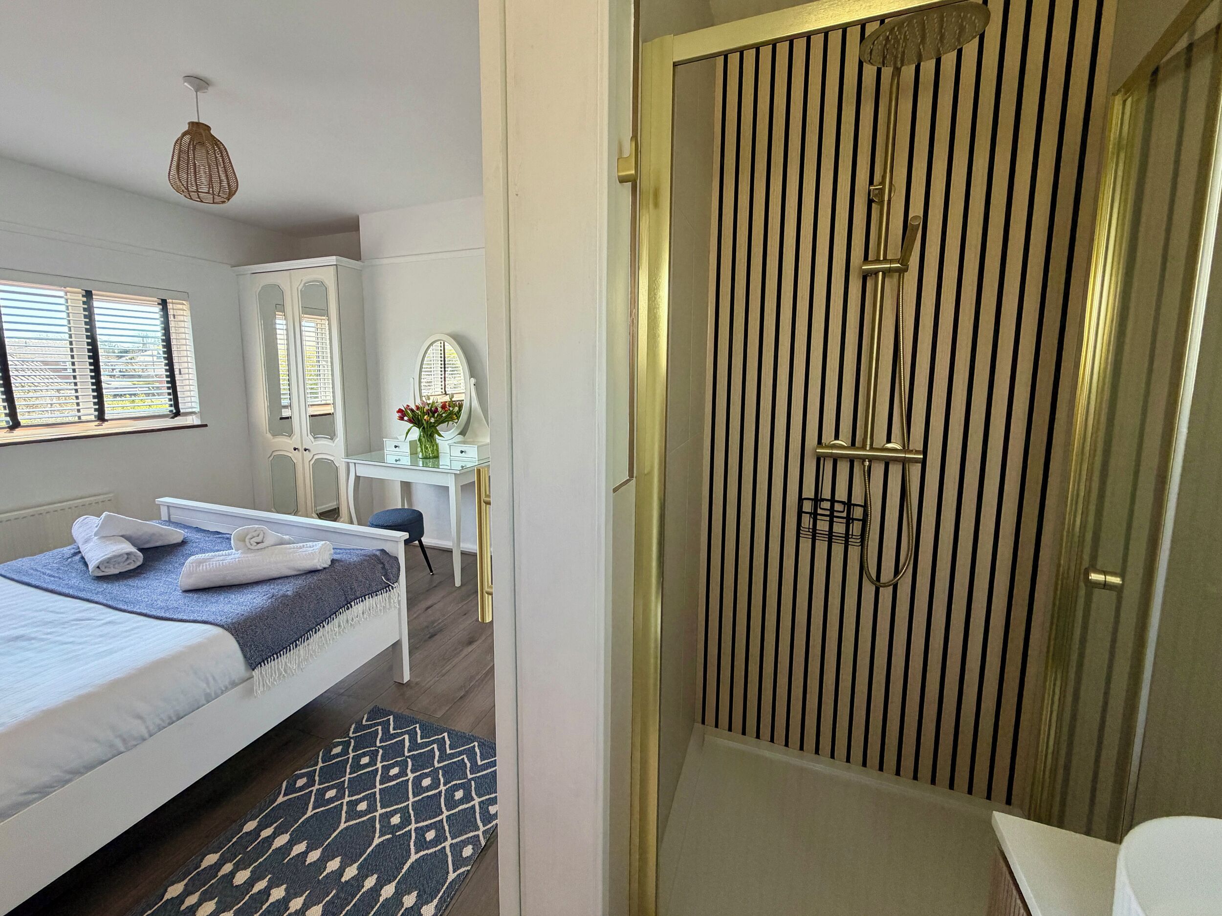 A bedroom with a bed, a vanity with a mirror, a desk, and a shower area with a vertical panel design.