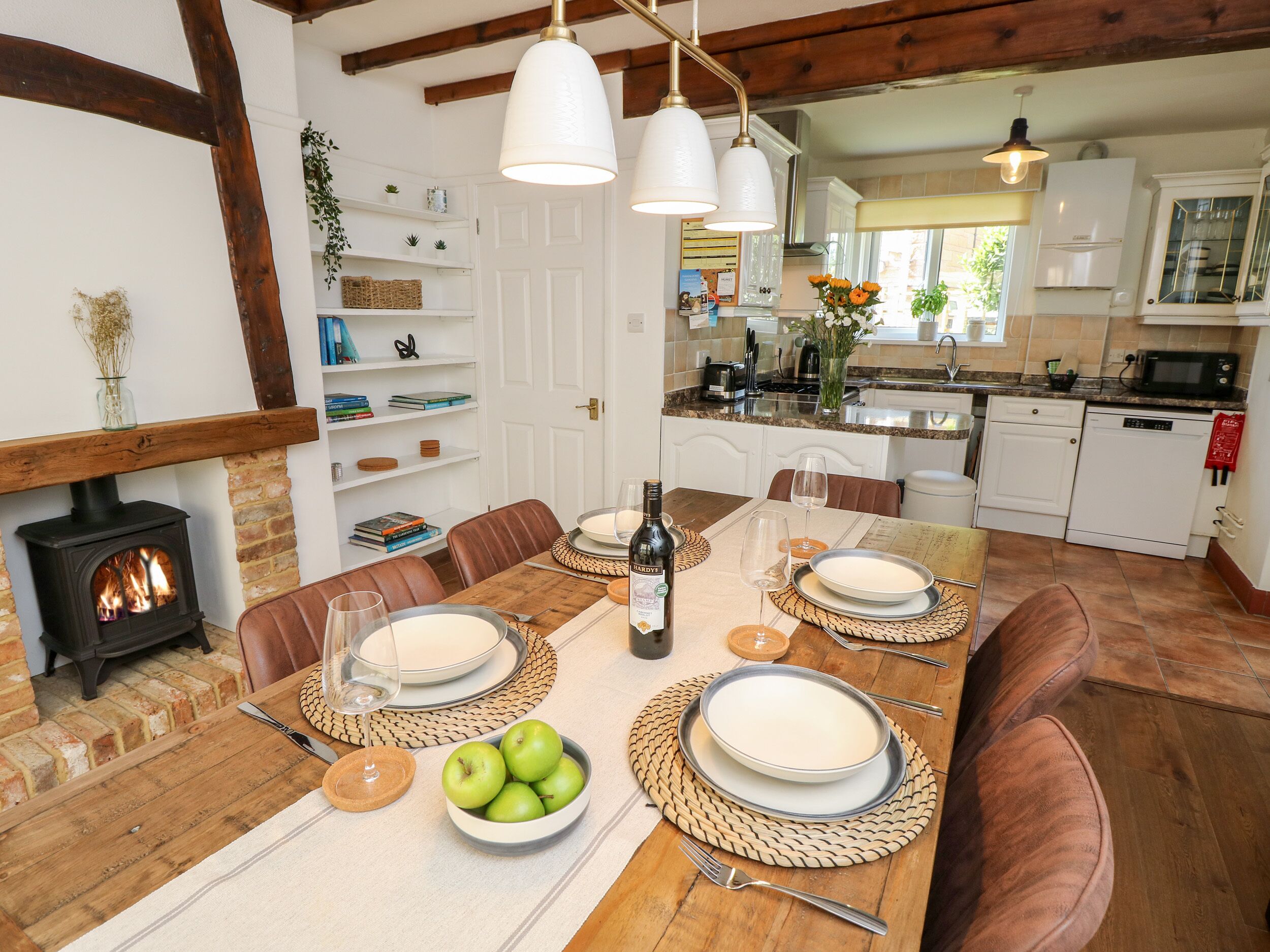 A dining area with a wooden table set for four, surrounded by brown leather chairs. A fireplace is visible to the left, and a kitchen with white cabinets and a stove is in the background.
