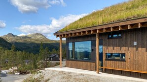 Un edificio moderno de madera con ventanas amplias y un techo cubierto de césped, rodeado de un paisaje montañoso.