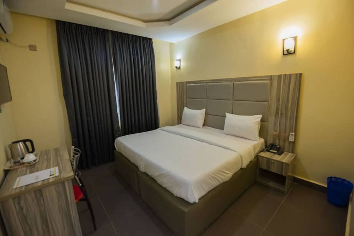 A hotel room with a large bed, a bedside table, a desk, a chair, a curtain, and a blue trash bin.