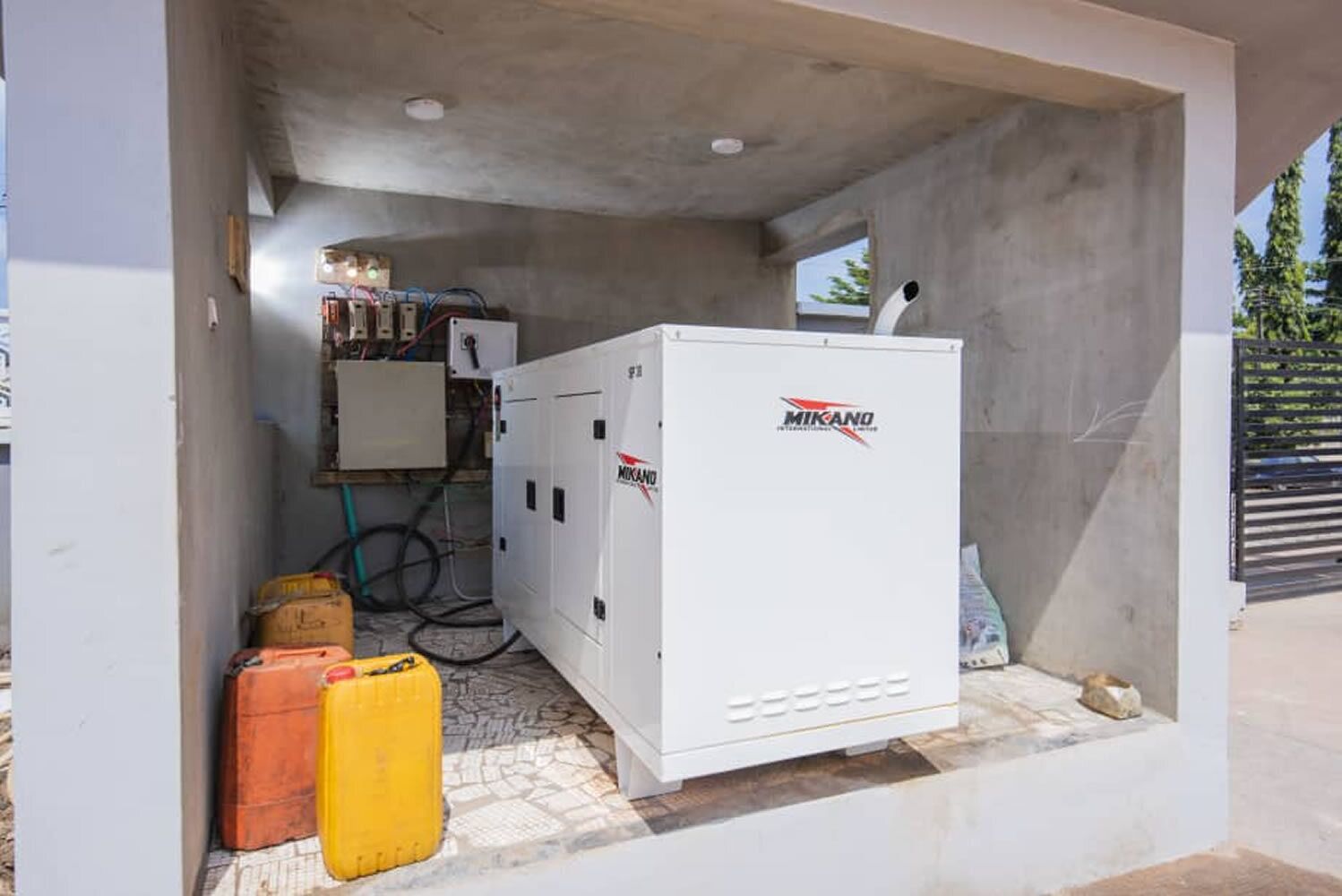 Un cuarto de servicio con un panel eléctrico blanco grande, identificado con la etiqueta "Mikano", y dos cilindros de gas amarillos.