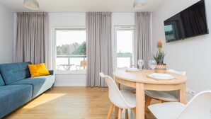 Een moderne woonkamer met een blauwe bank, een eettafel met stoelen en een aan de muur bevestigde televisie.