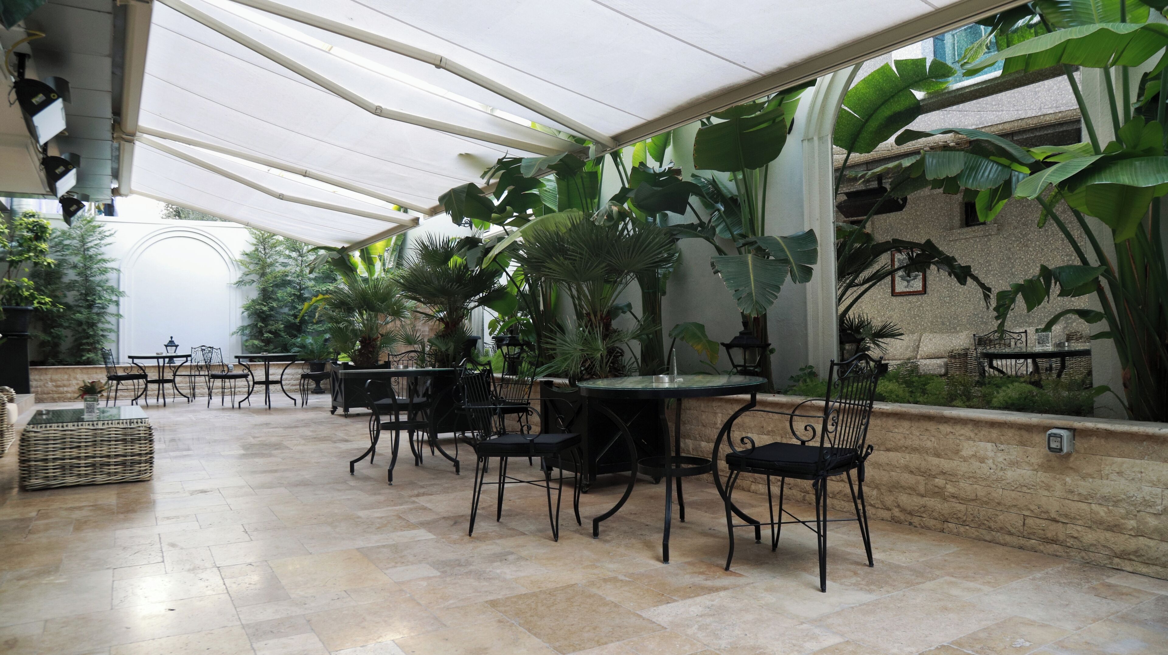 An indoor patio area with tiled flooring, black metal chairs, and tables. There are large green plants and a stone wall in the background.
