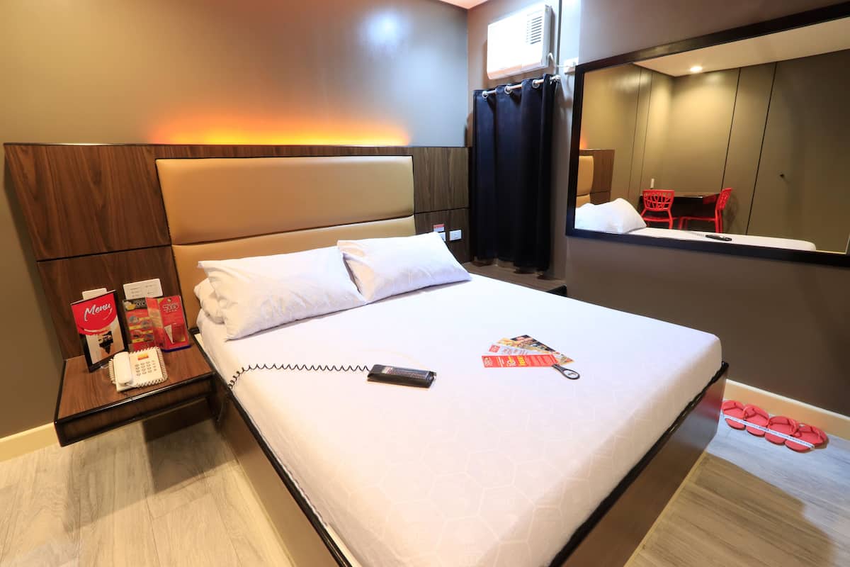 A hotel room with a large bed, a bedside table with a phone and brochures, a mirror, and a small table with red chairs.