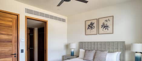A hotel room with a wooden door, a bed with a grey headboard, two bedside tables with lamps, a tray with a cup and a book, and two framed botanical prints on the wall.