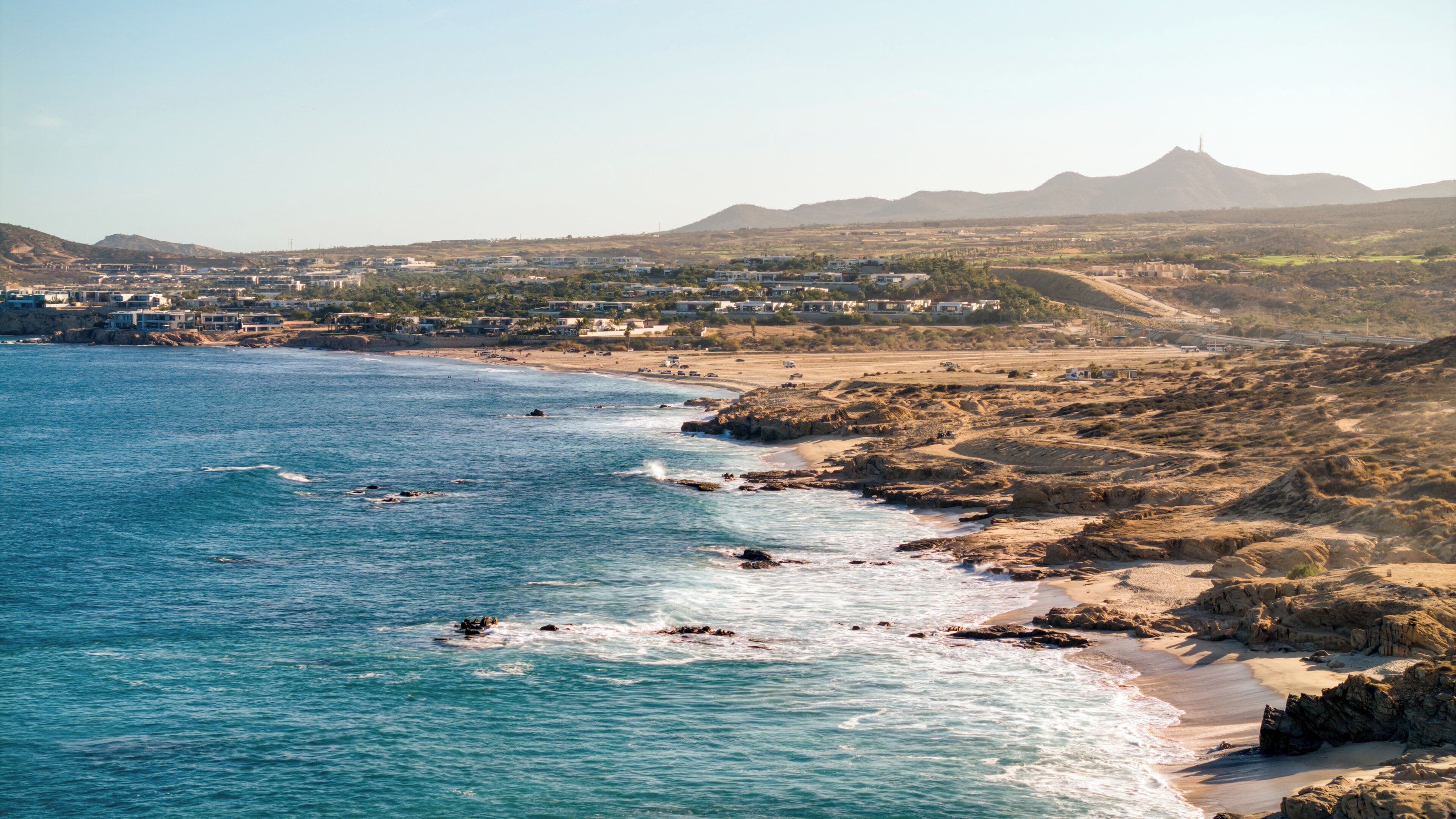 Un pueblo costero con playa, costa rocosa y montañas a lo lejos.