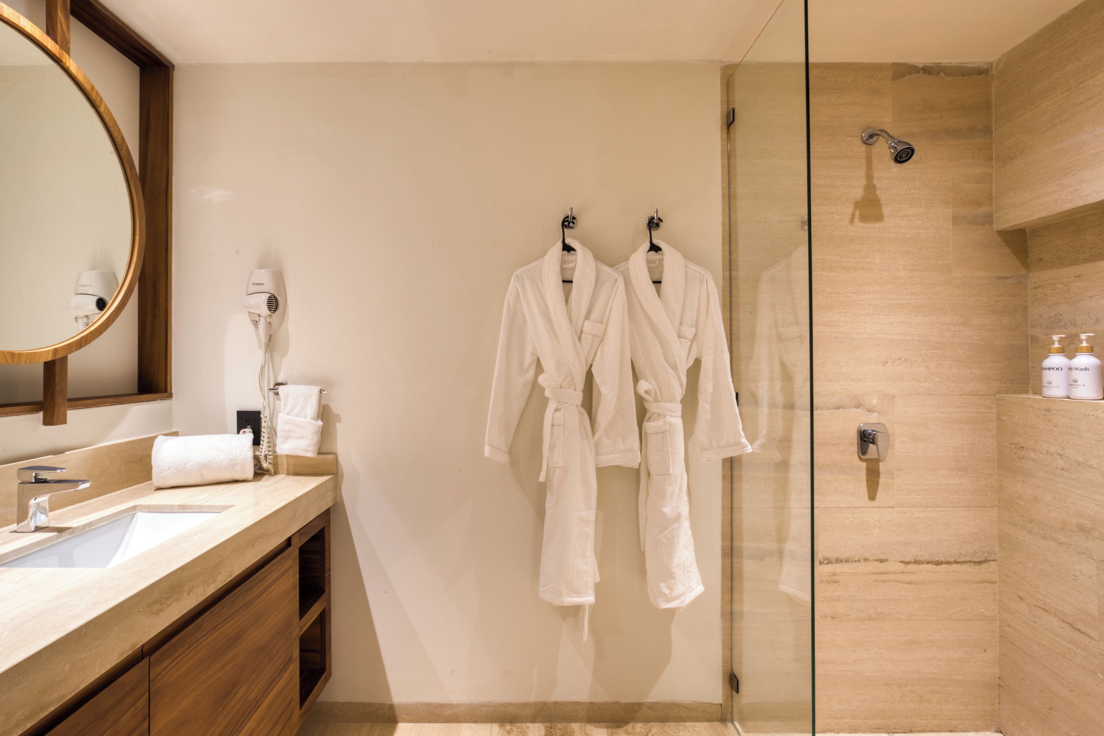 A modern bathroom with a glass shower enclosure, a wooden vanity, and two white robes hanging on hooks.