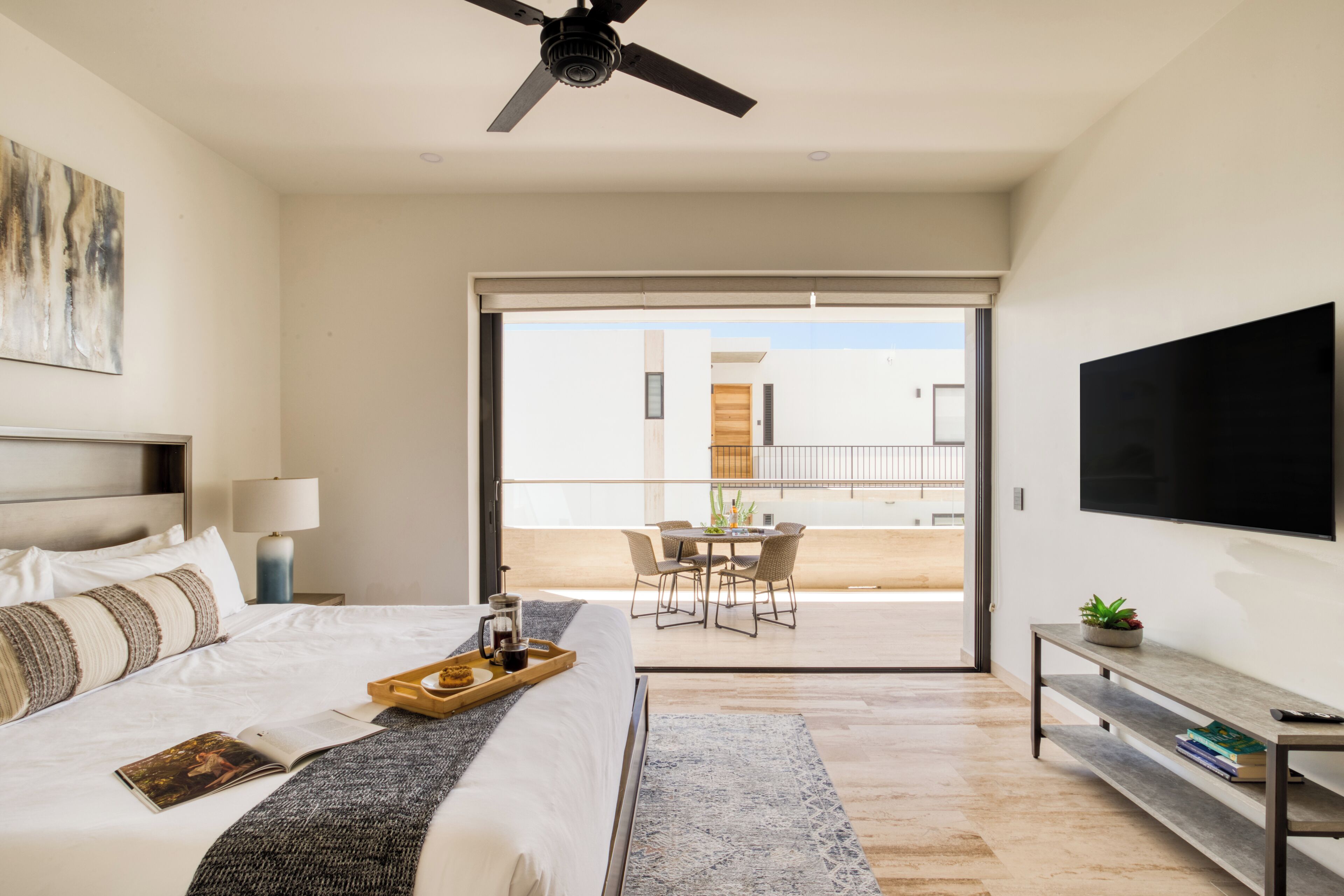 A modern hotel room with a bed, a coffee table with a tray, a TV, and a balcony with a dining area.
