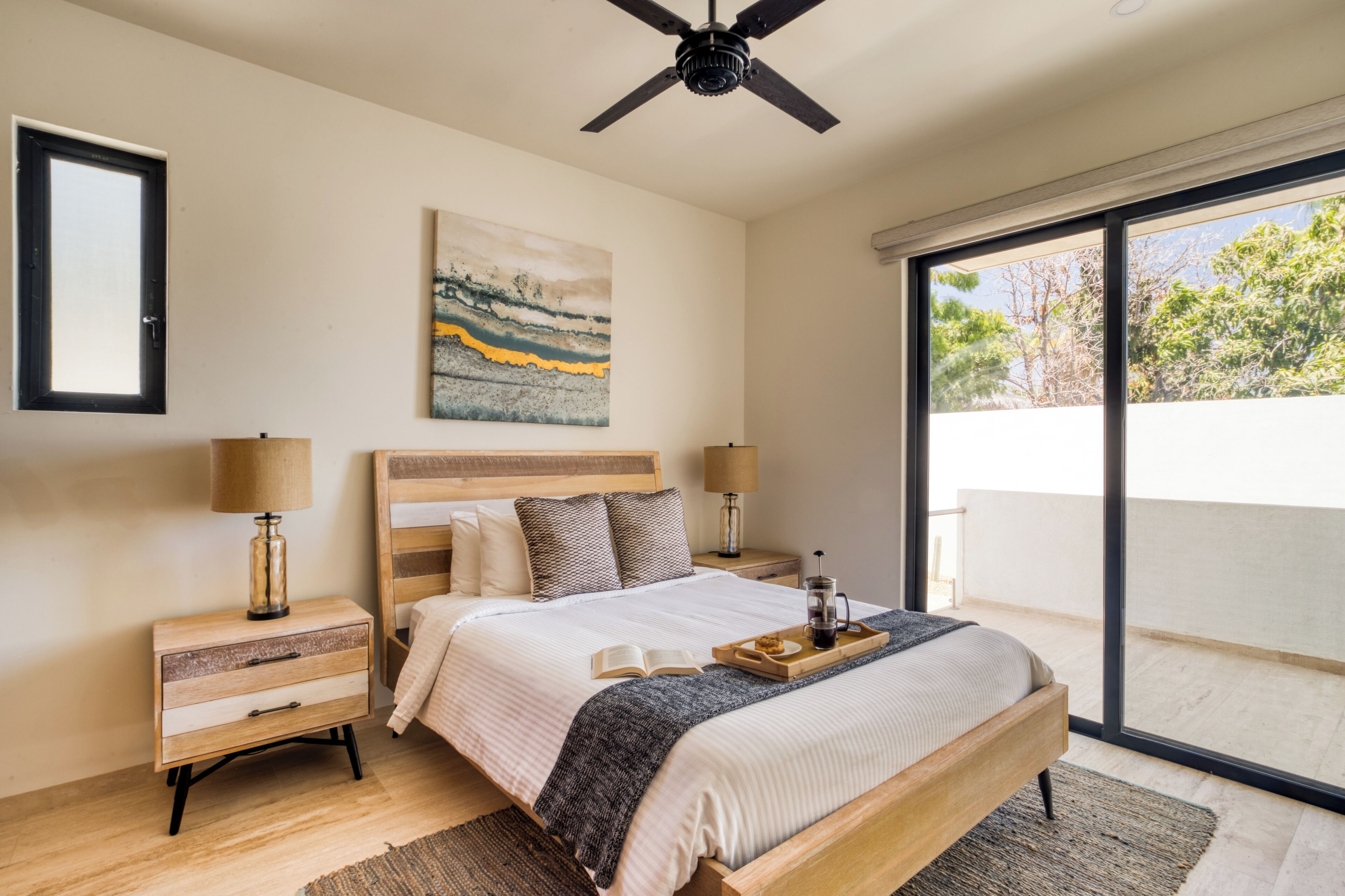 A bedroom with a large bed, wooden headboard, two bedside tables with lamps, a window with a view of trees, and a ceiling fan.