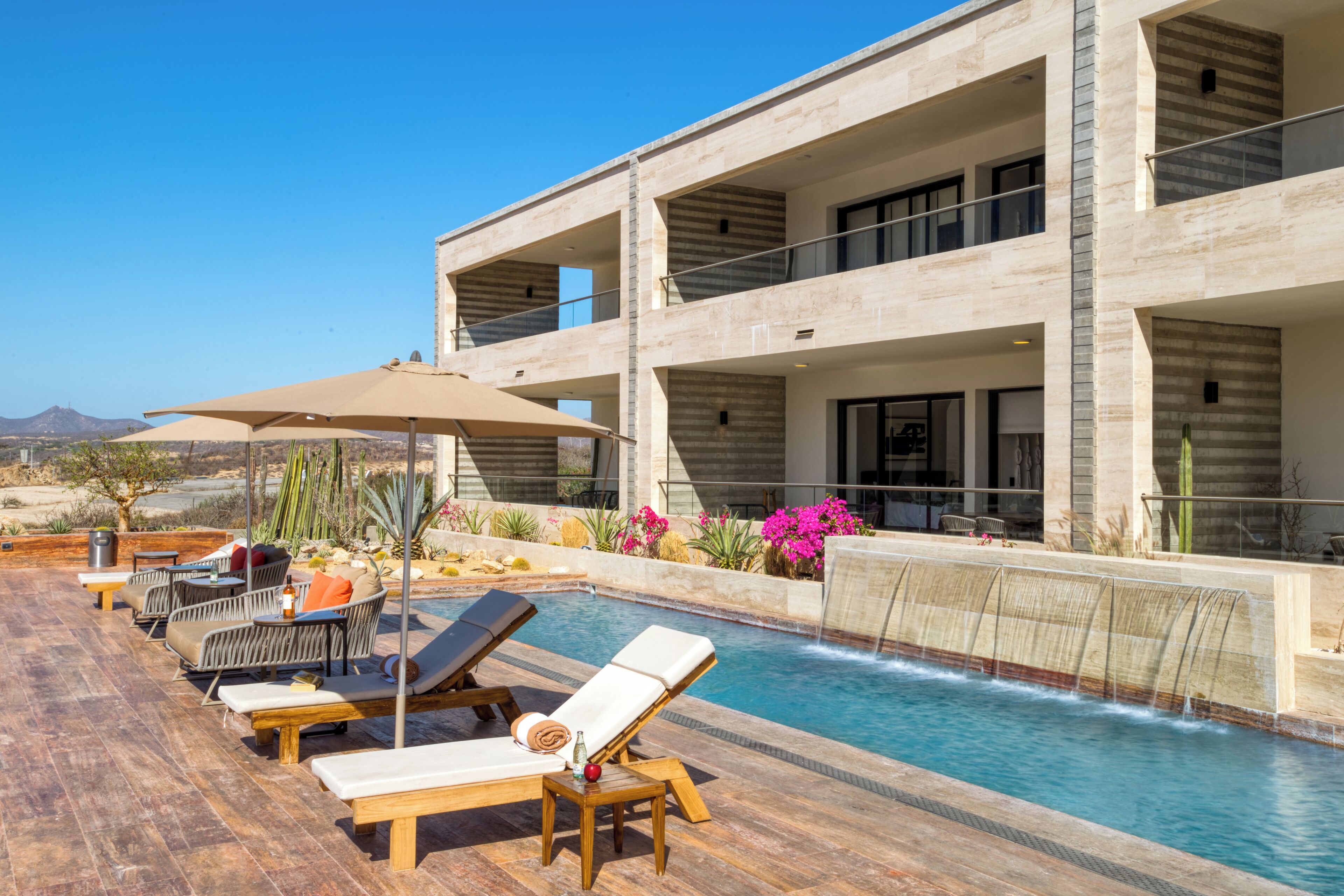 A modern poolside area with lounge chairs and a table, a pool with a waterfall feature, and a view of the surrounding landscape.
