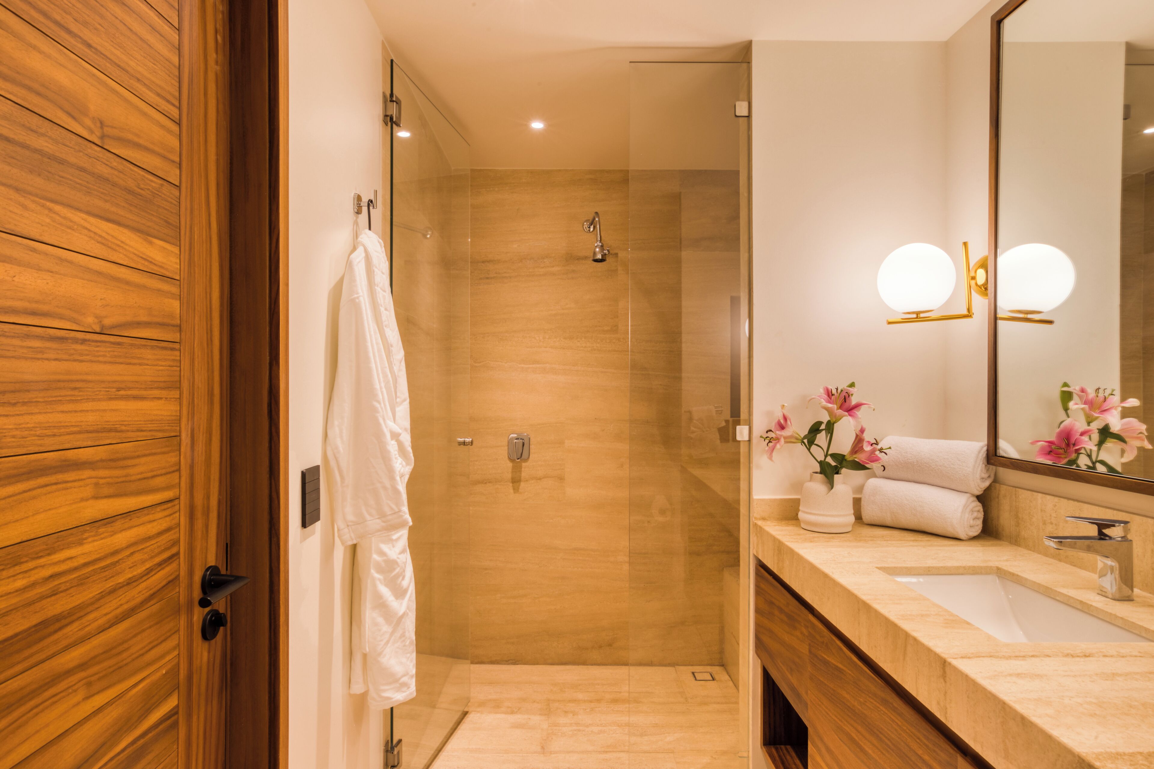 Ein modernes Badezimmer mit Holztür, Duschkabine aus Glas, Waschtisch und Spiegel mit runden Leuchten.
