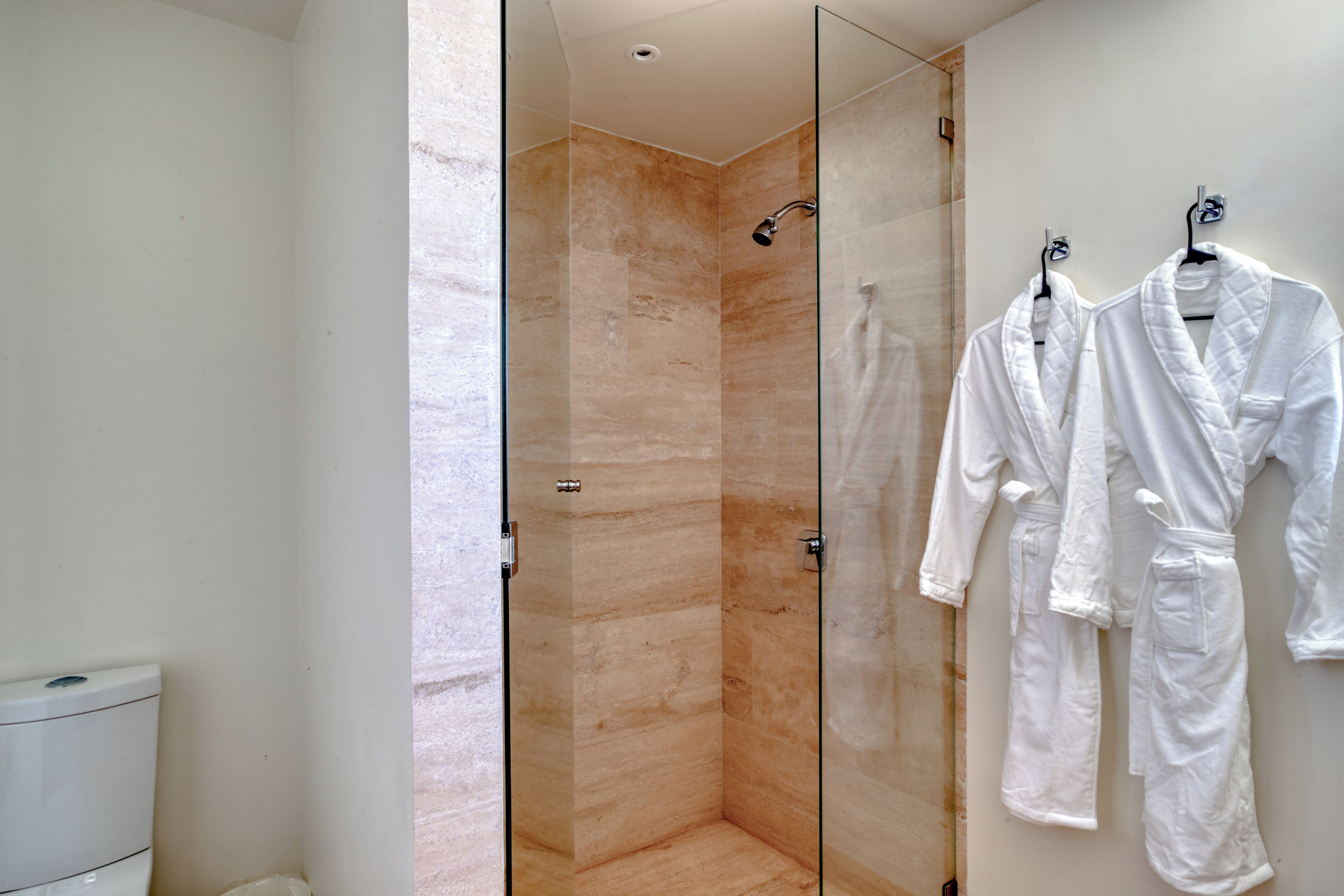 A modern bathroom with a glass-enclosed shower, white tiled walls, and two white robes hanging on hooks.