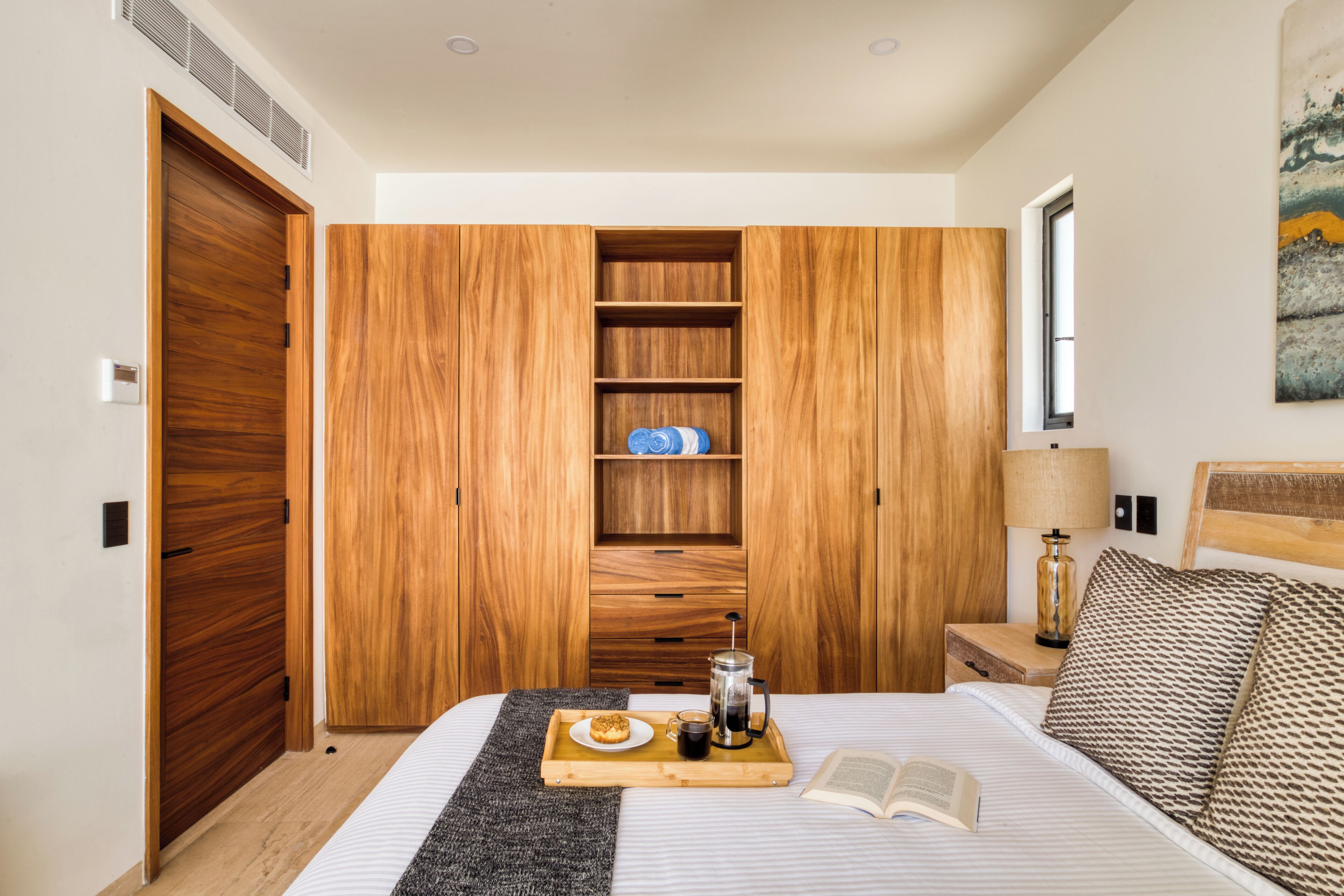 A bedroom with a wooden wardrobe, a bedside table with a lamp, a tray with breakfast, and an open book on the bed.