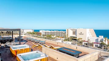 A rooftop terrace with multiple hot tubs, outdoor seating, and a view of the ocean and cityscape.