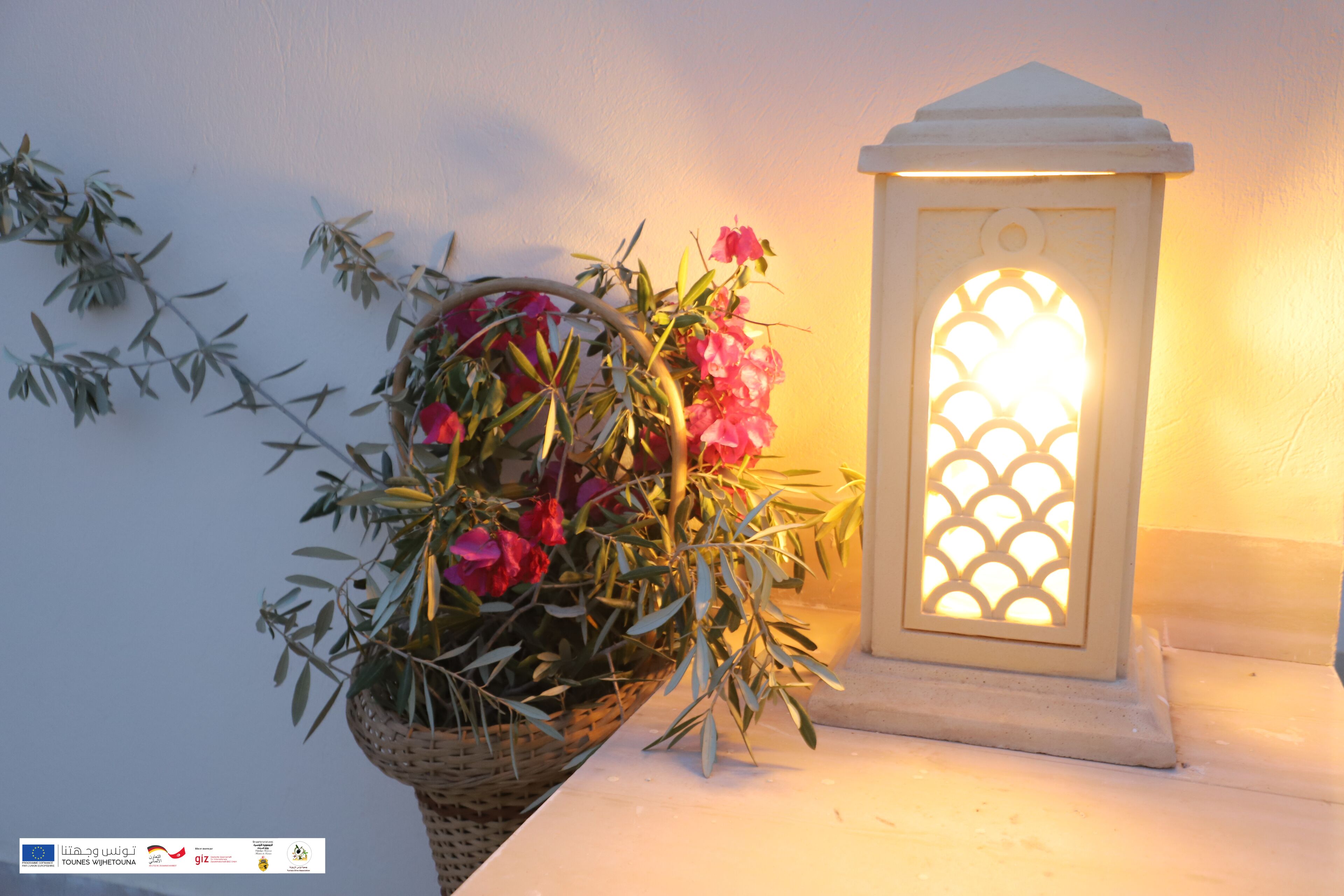 Un farol blanco con un diseño decorativo en la ventana, junto a una cesta de mimbre con flores rosadas y follaje verde.