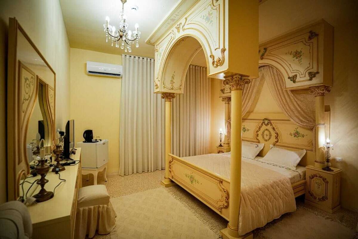 Uma cama com dossel e detalhes ornamentados, uma mesa de cabeceira com abajur e um espelho com cortina pendurada.