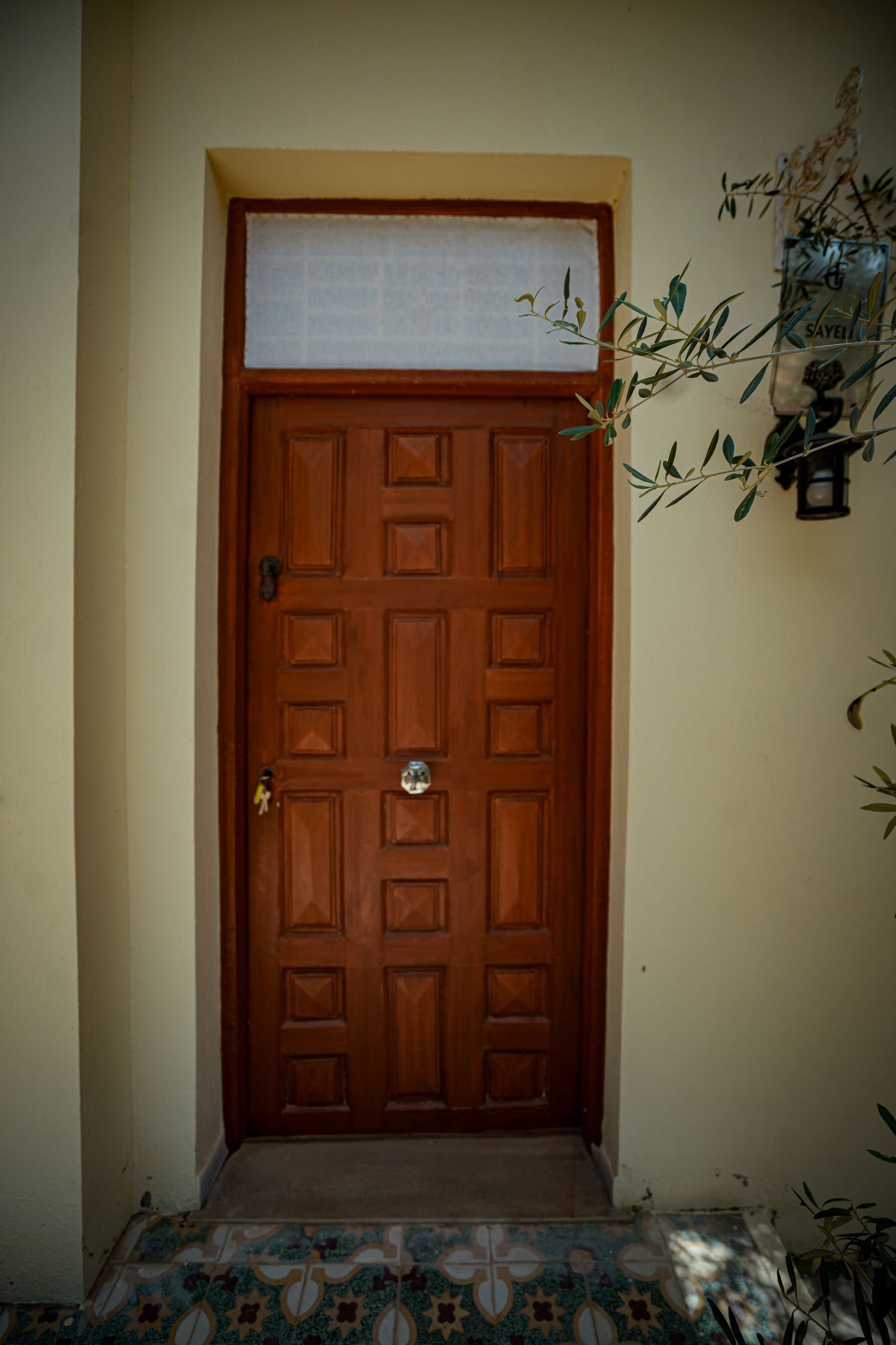 A wooden door with a metal handle and a keyhole, set in a hallway with patterned floor tiles.