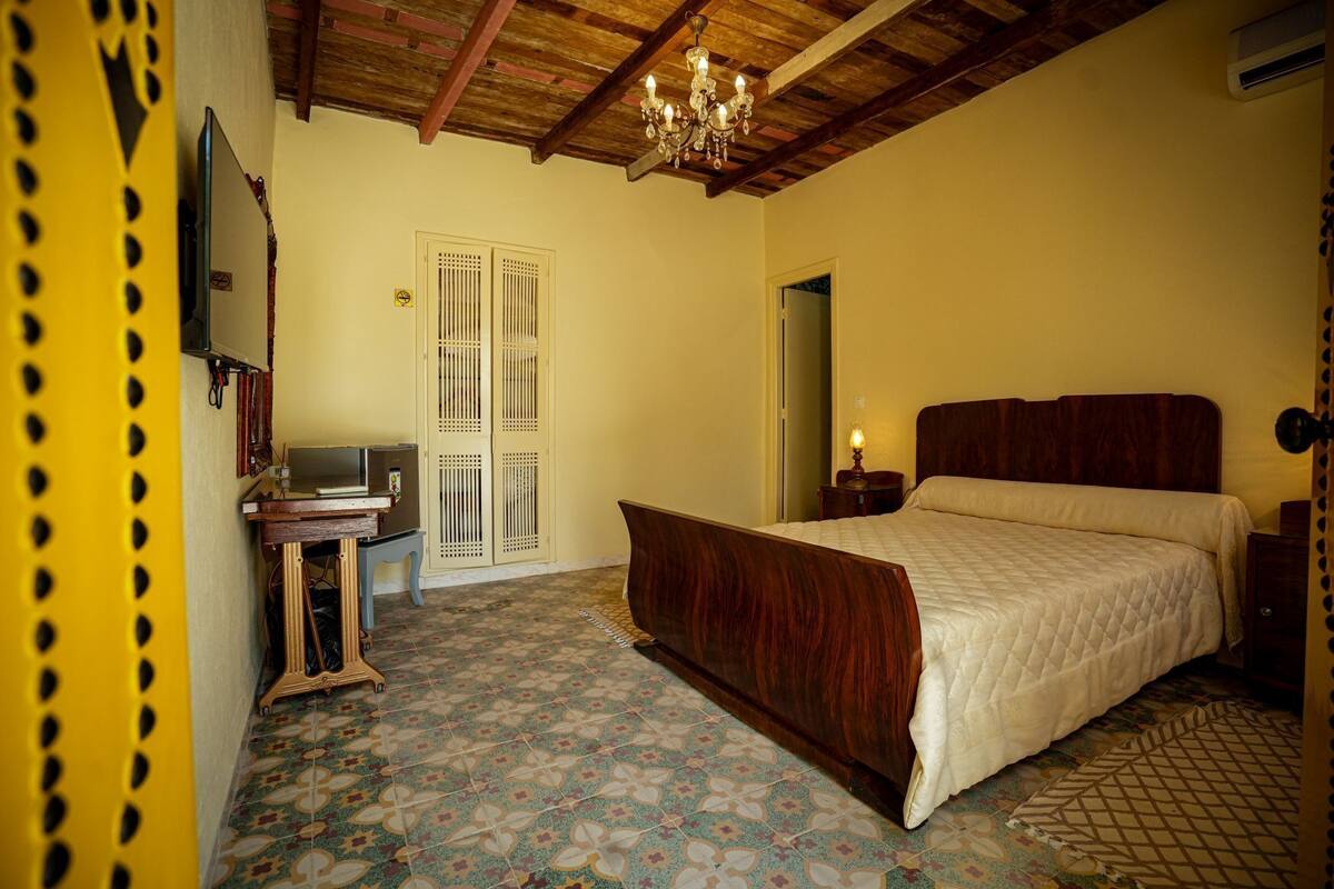 Quarto com cama de madeira, escrivaninha com cadeira, candelabro e ladrilhos estampados.
