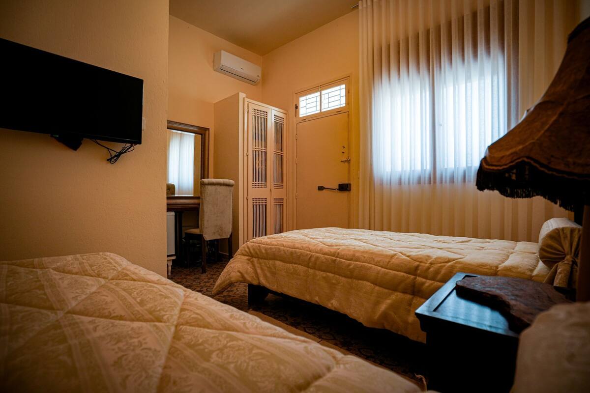 Quarto de hotel com duas camas, televisão fixada na parede, guarda-roupa e janela com cortinas.