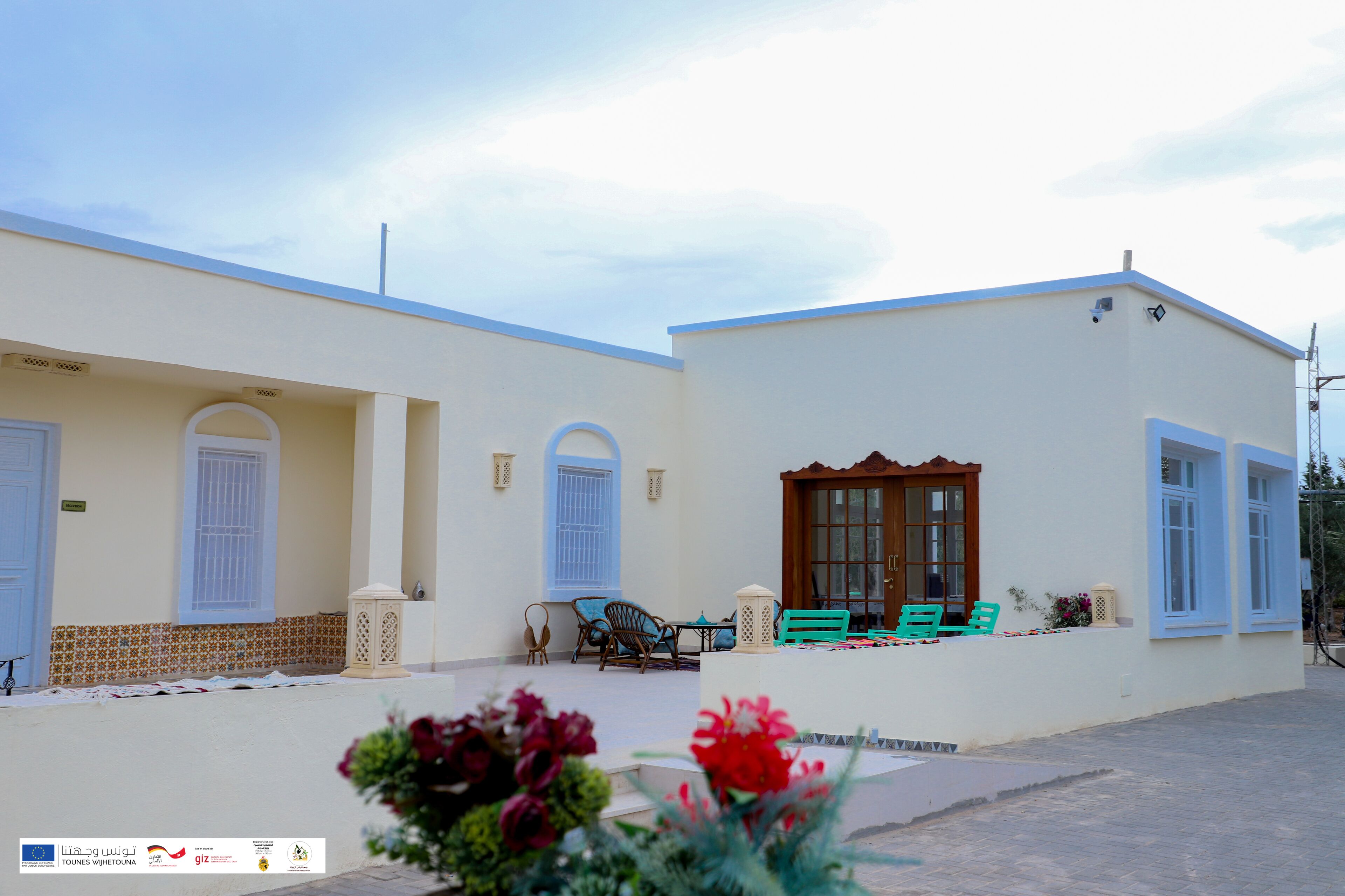 A two-story building with a flat roof, white exterior walls, and blue trim. There are two windows with white shutters, a door, and a small porch area with a bench and a table.