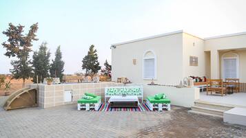 A patio area with a white table and two green cushioned chairs, a white bench, and a striped rug.