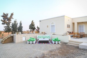 A patio area with a white table and two green cushioned chairs, a white bench, and a striped rug.