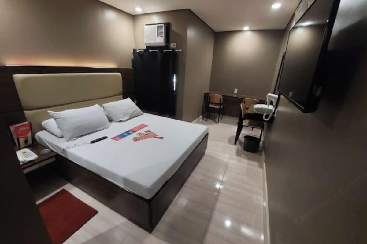 A modern hotel room with a large bed, a desk, a chair, a TV, and an air conditioning unit.