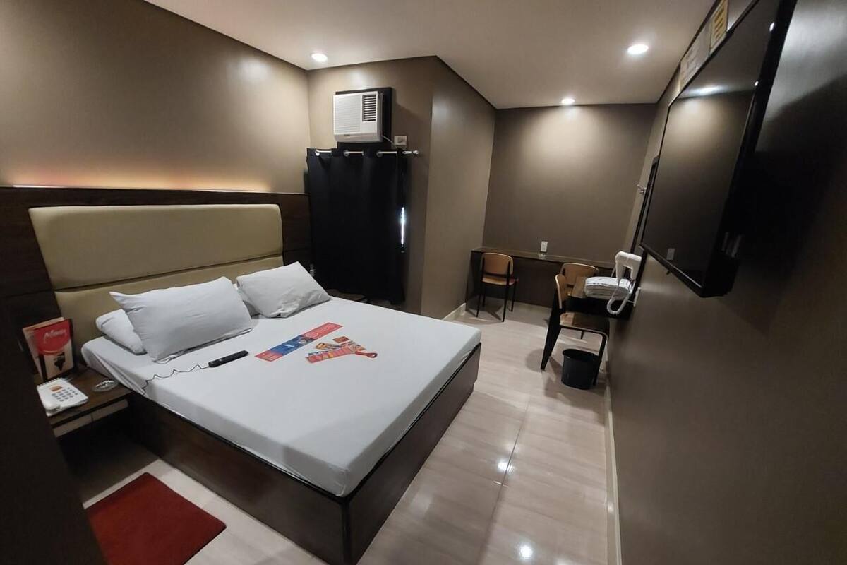 A modern hotel room with a large bed, a desk, a chair, a TV, and a minibar.
