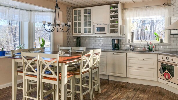 A kitchen with white cabinets, a wooden dining table, and chairs. There is a window with a view of trees outside.