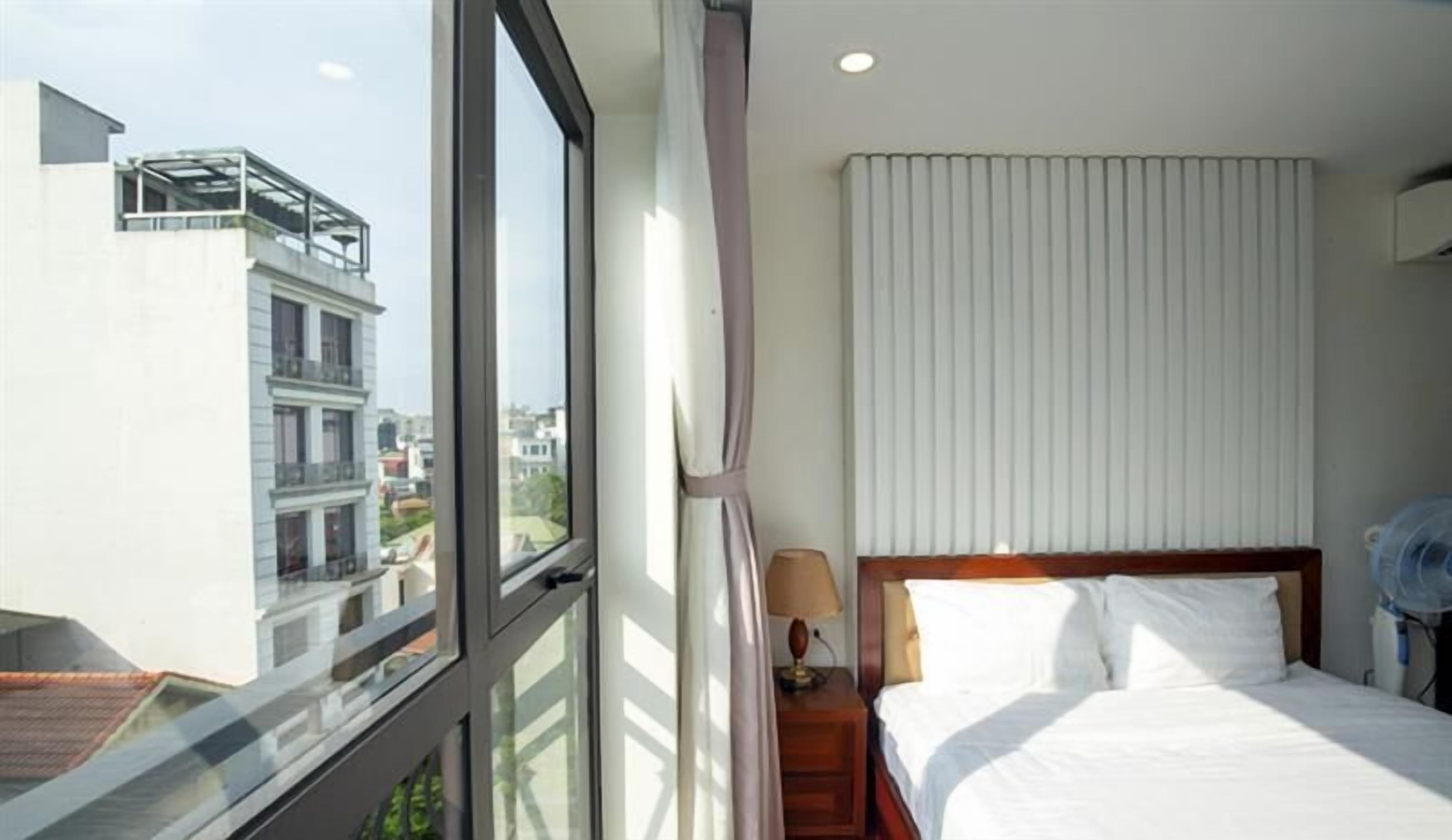 A hotel room with a large window, a bed with white linens, a nightstand with a lamp, and a view of buildings outside.