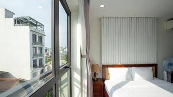 A hotel room with a large window, a bed with white linens, a nightstand with a lamp, and a view of buildings outside.