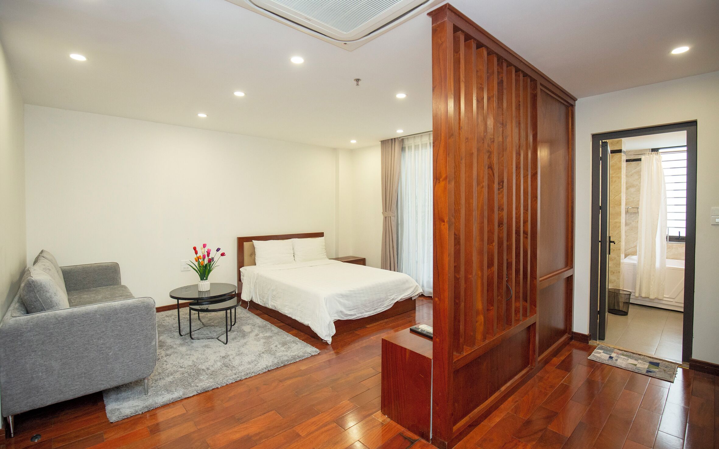A modern hotel room with a bed, a gray sofa, a small table with a vase of flowers, and a wooden partition.