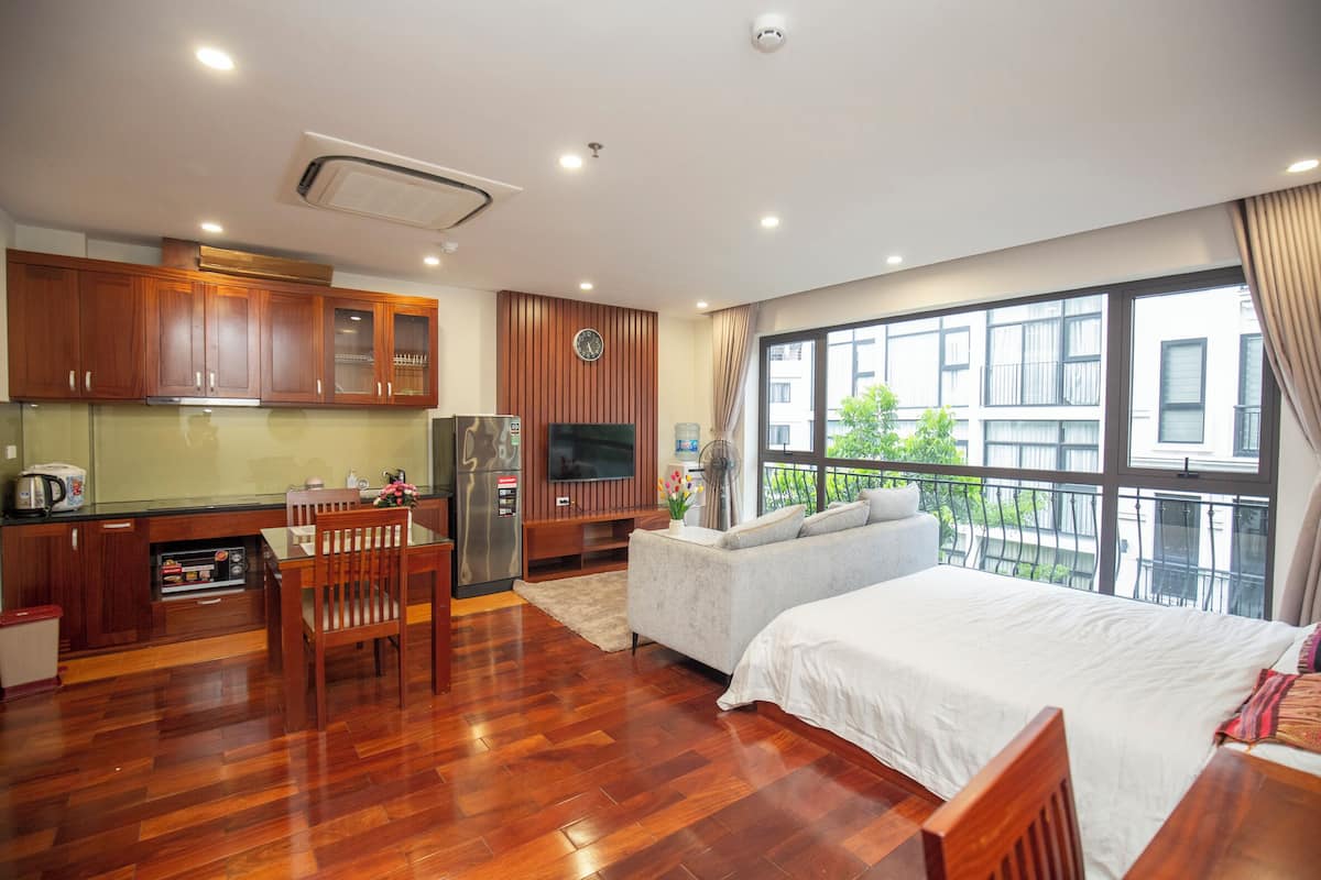 Habitación de hotel moderna con piso de madera, una cama grande, un sofá, un televisor, una zona de cocina con gabinetes de madera y un balcón con vistas.