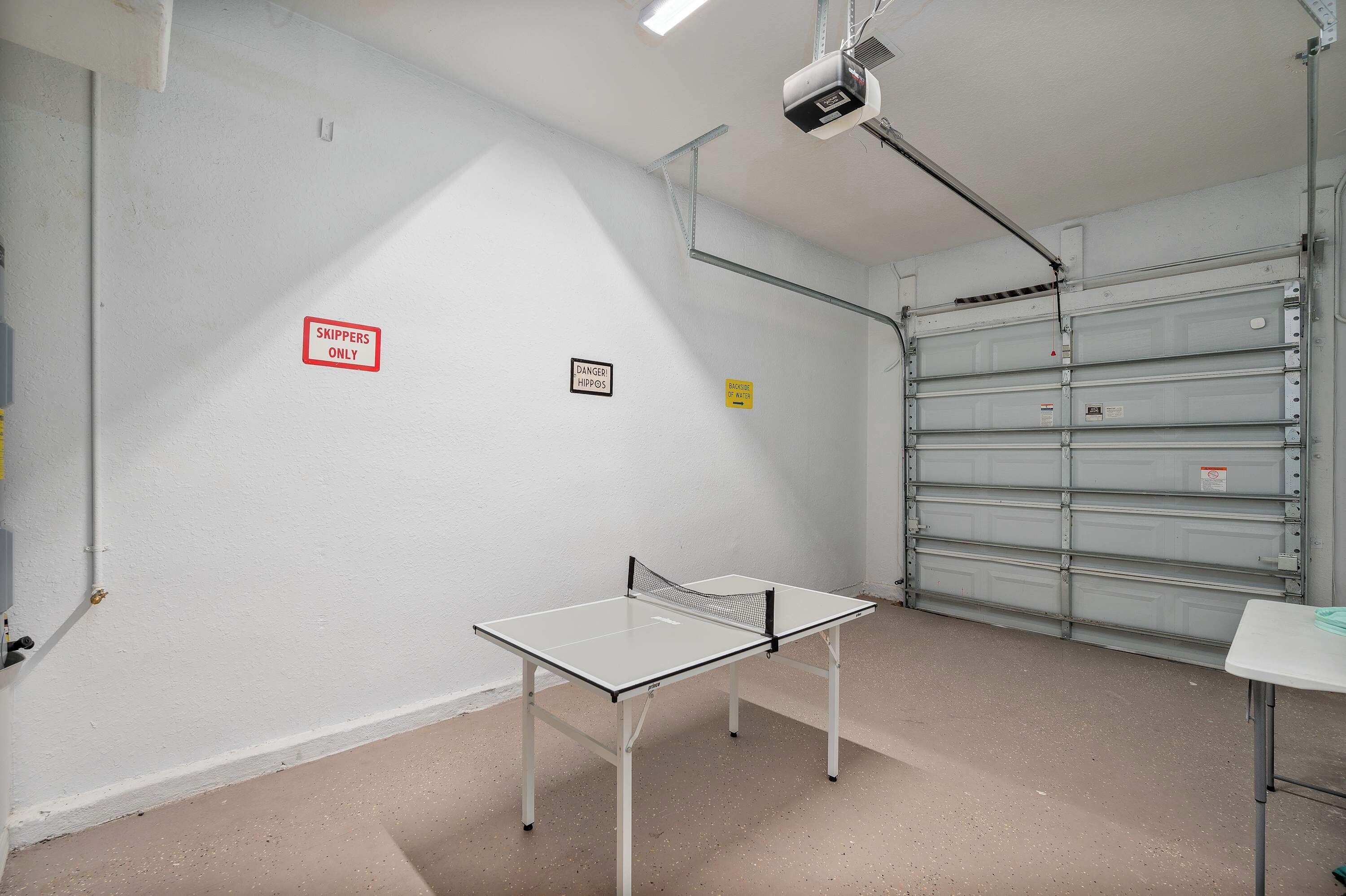 Una mesa de ping pong en una habitación con paredes blancas, un proyector montado en el techo y una puerta de garaje cerrada.