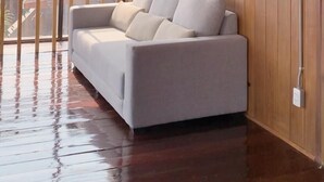 A modern room with a wooden paneled wall, a white sofa with cushions, a ceiling fan, and a large window offering a view of buildings outside. - U Villa Chiangkhan (Chiang Khan)