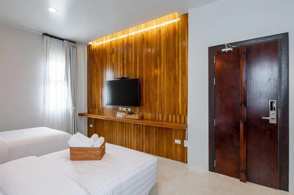 A hotel room with two beds, a television mounted on a wooden panel wall, a dark wooden door, and a basket of toiletries on one bed.