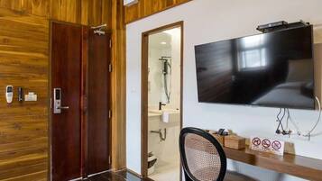 Habitación de hotel con pared de madera, un televisor montado en la pared, un escritorio con silla y un baño visible a través de una puerta de cristal.