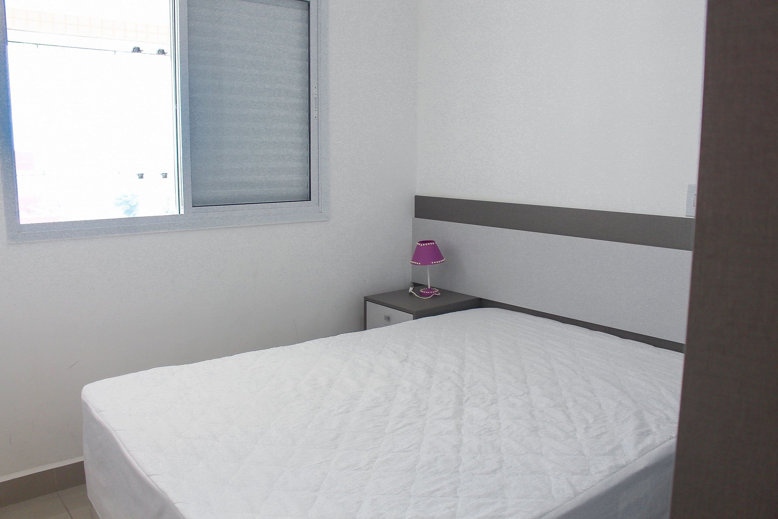 Ein kleines, ordentlich bezogenes Bett in einem Zimmer mit Fenster, einem Nachttisch mit Lampe und einer schlichten Wand.