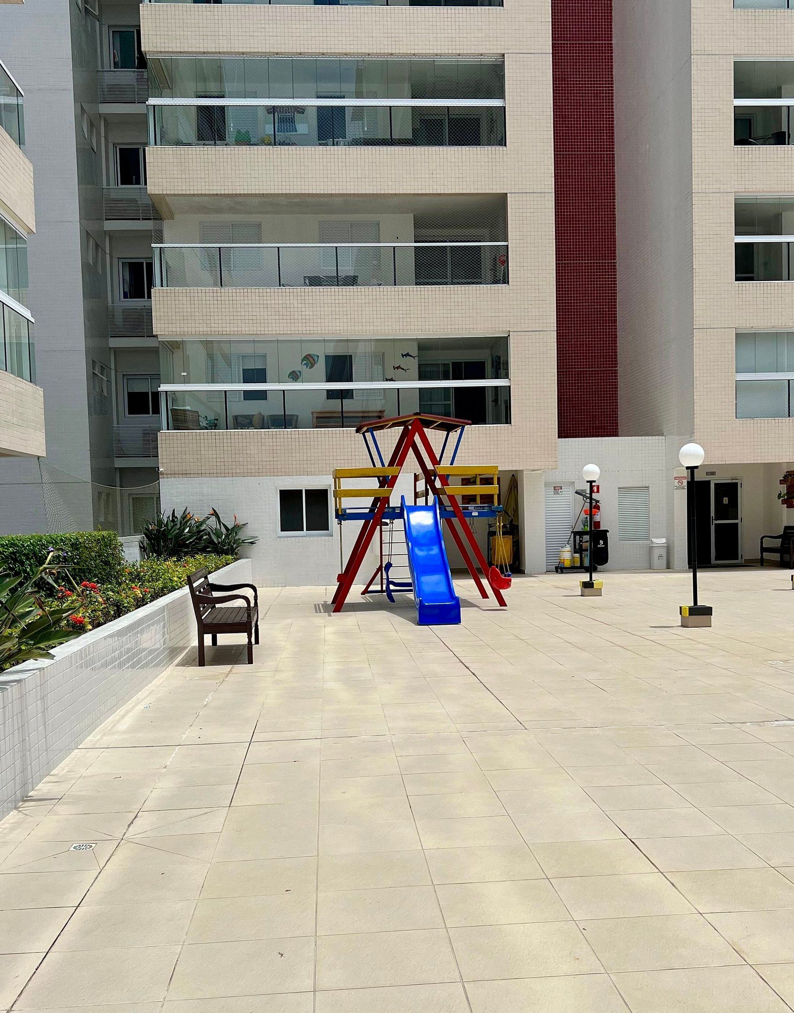 A playground with a slide and swings, surrounded by a building with multiple balconies and a tall red column.